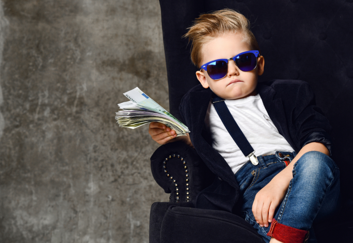 un enfant tenant une liasse de billet assis dans un fauteuil trop grand avec des lunettes de soleil et des bretelles