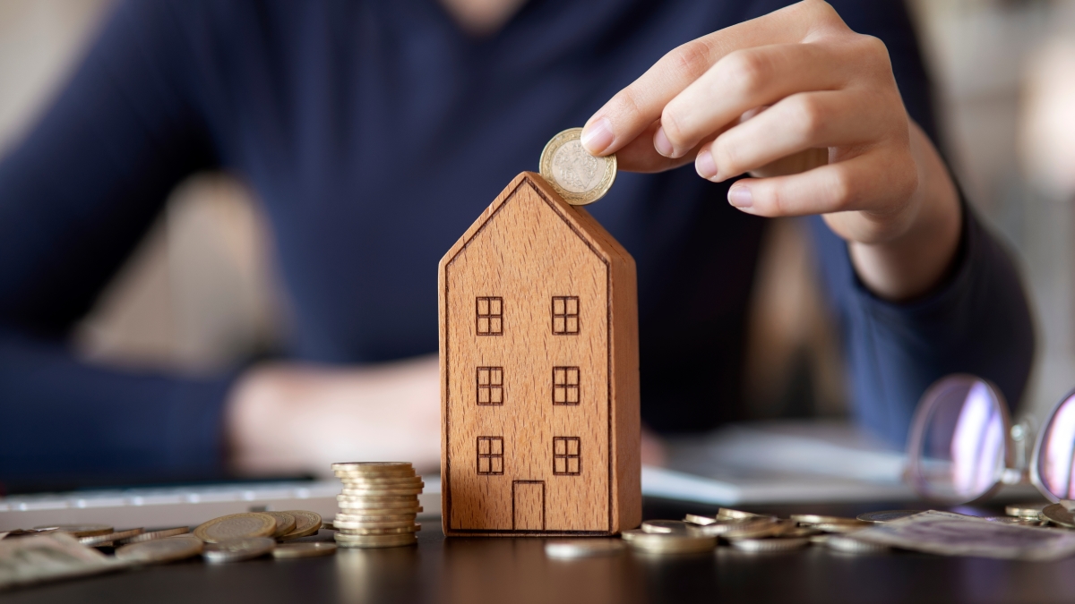 Loi SRU Immobilier — Une personne insère des pièces de monnaie dans une maison miniature